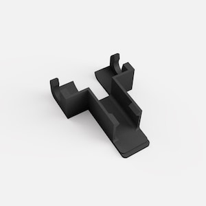 Puede incluir: Un componente de plástico negro con un diseño angular único. La pieza presenta una base rectangular, un canal central y dos secciones curvas elevadas. El objeto está sobre un fondo blanco liso.