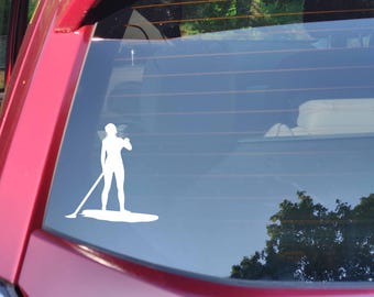 May include: White vinyl decal of a person paddleboarding on a red car window. The silhouette shows a person standing on a paddleboard, holding a paddle. The car is a deep red color.