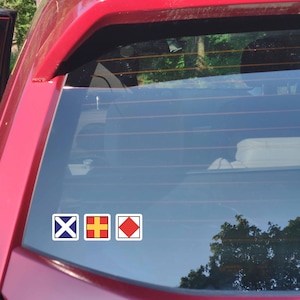 May include: Three nautical signal flag stickers on a car window. The flags are in white squares with blue, yellow, and red designs. The car is red, and the background shows trees and sky.