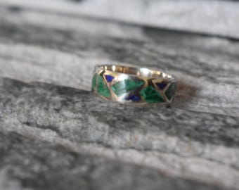 May include: A silver ring with a geometric design featuring green and blue inlays. The ring has a band with a gold-colored interior. The ring is displayed on a gray, textured surface.