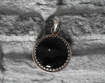 May include: A round, black gemstone pendant with a silver-colored bail. The gemstone is set in a decorative, silver-colored bezel with a rope-like design. The pendant is displayed against a textured, gray background.