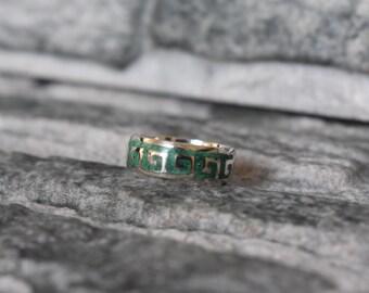 May include: A silver ring with a repeating Greek key design filled with green inlay. The ring is set against a textured gray background, showcasing the intricate details of the jewelry.