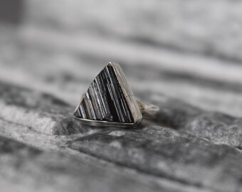Puede incluir: Un anillo de plata con una piedra triangular negra y gris. La piedra tiene un patrón de rayas y está engastada en una banda de plata. El anillo se muestra sobre una superficie gris texturizada.