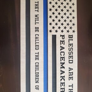 Peut inclure: Une pancarte en bois avec un design de drapeau américain bleu et blanc. Le texte "Blessed are the peacemakers for they will be called the children of God" est écrit sur la pancarte.
