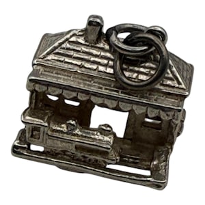 May include: A silver-toned charm in the shape of a small house with a detailed roof, windows, and a train. The charm has a loop at the top for attachment to a bracelet or necklace. The charm is made of metal and has a vintage appearance.