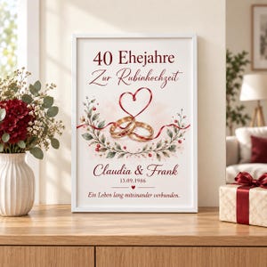 May include: A framed vertical print featuring the text 40 Ehejahre Zur Rubinhochzeit, Claudia & Frank, 13.09.1986, and Ein Leben lang miteinander verbunden. The design includes two gold rings, a red ribbon heart, and floral greenery.