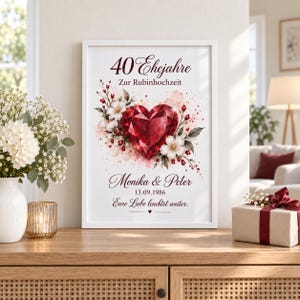 Op de afbeelding: Een ingelijste verticale print met een gefacetteerde rode hartafbeelding omringd door witte bloemen en rode bessen. De tekst luidt 40 Ehejahre Zur Rubinhochzeit, Monika & Peter, 13.09.1986 en Eure Liebe leuchtet weiter, met een klein hartsymbool.