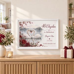 Op de afbeelding: Een ingelijste horizontale print met twee witte zwanen op een meer die een hartvorm vormen met hun nekken, omringd door donkerrode rozen. De tekst luidt 40 Ehejahre, Zur Rubinhochzeit, Birgit & Uwe, 13.09.1986, Für all die schönen Jahre vor euch.