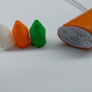 Könnte beinhalten: Ein orangefarbener und weißer elektrischer Farbsprüher mit drei farbigen Düsen: weiß, orange und grün. Der Sprüher hat eine weiße Basis mit einem Knopf und eine Reihe konzentrischer Kreise.