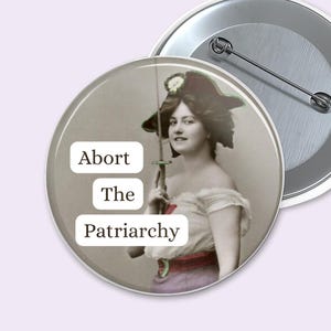 Può includere: Un bottone rotondo con un'immagine vintage di una donna che tiene una spada, con il testo "Abort The Patriarchy" in bianco. Il bottone è grigio chiaro con un retro a spillo argentato. Un secondo bottone è visibile.