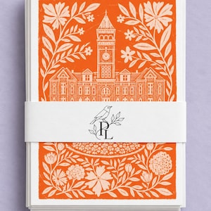Könnte beinhalten: Ein Stapel rechteckiger Karten, zusammengehalten von einem weißen Papierband mit einer schwarzen Strichzeichnung eines Vogels auf den Buchstaben R und L. Die Karten zeigen eine orangefarbene und cremefarbene Illustration eines Uhrturms.