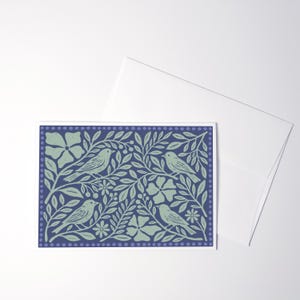 Puede incluir: Una tarjeta de felicitaci&oacute;n rectangular con fondo azul oscuro y un patr&oacute;n de impresi&oacute;n en bloque verde claro de cuatro p&aacute;jaros posados entre hojas, flores y bayas estilizadas, enmarcado por un borde de puntos peque&ntilde;os, junto a un sobre blanco.