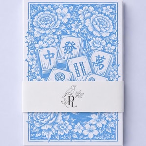 Könnte beinhalten: Ein Stapel rechteckiger Karten mit einem blau-weißen Blumenmuster und Illustrationen von Mahjong-Steinen. Ein weißes Papierband umschließt die Mitte und zeigt eine schwarze Strichzeichnung eines Vogels auf den Buchstaben R und L.