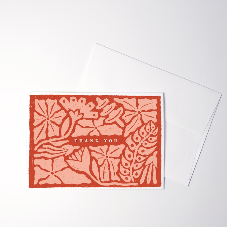 K&ouml;nnte beinhalten: Eine rechteckige Gru&szlig;karte mit einem floralen Muster im Blockdruck-Stil in Terrakotta- und Hellrosat&ouml;nen. Der Text THANK YOU ist in Wei&szlig; in der Mitte der Karte gedruckt. Ein schlichter wei&szlig;er Umschlag befindet sich hinter der Karte.
