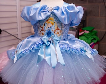 Puede incluir: Un vestido de tul tutu azul claro con un corpiño de satén con un estampado de Cenicienta y pedrería. El vestido tiene un lazo de satén azul y un ribete de volantes de satén azul claro.