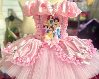 Puede incluir: Un vestido de tul rosa con una falda de tutú y un corpiño de satén con un estampado de princesas Disney. El corpiño tiene un lazo de satén rosa con un strass en el centro. El vestido tiene volantes de satén rosa alrededor del dobladillo.
