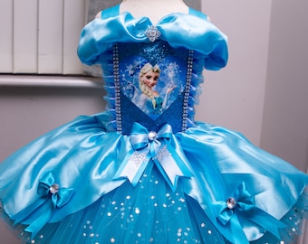 Puede incluir: Un vestido de tul azul con una falda de tul y un corpiño de satén con un diseño de corazón brillante con una imagen de Elsa de Frozen. El vestido tiene un lazo de satén azul en la parte delantera y un lazo de satén blanco en la parte posterior.