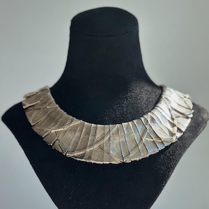May include: A wide, silver-toned collar necklace featuring a series of overlapping, fan-shaped metal segments with a brushed, textured finish. The necklace has a curved, rigid structure that sits flat against the neck, creating a layered appearance.