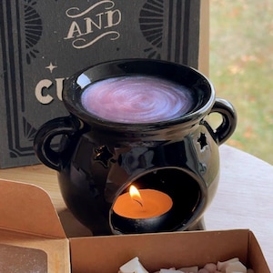 May include: A black ceramic oil burner with star-shaped cutouts, a lit candle, and a box of wax melts. The burner contains a purple liquid and is next to a book that says "Spells and". The wax melts are pastel-coloured.