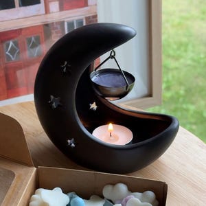 May include: A black crescent moon oil burner with star cutouts and a hanging dish. A lit candle is inside the moon, and a box of cloud-shaped wax melts in pastel colours is in the foreground.