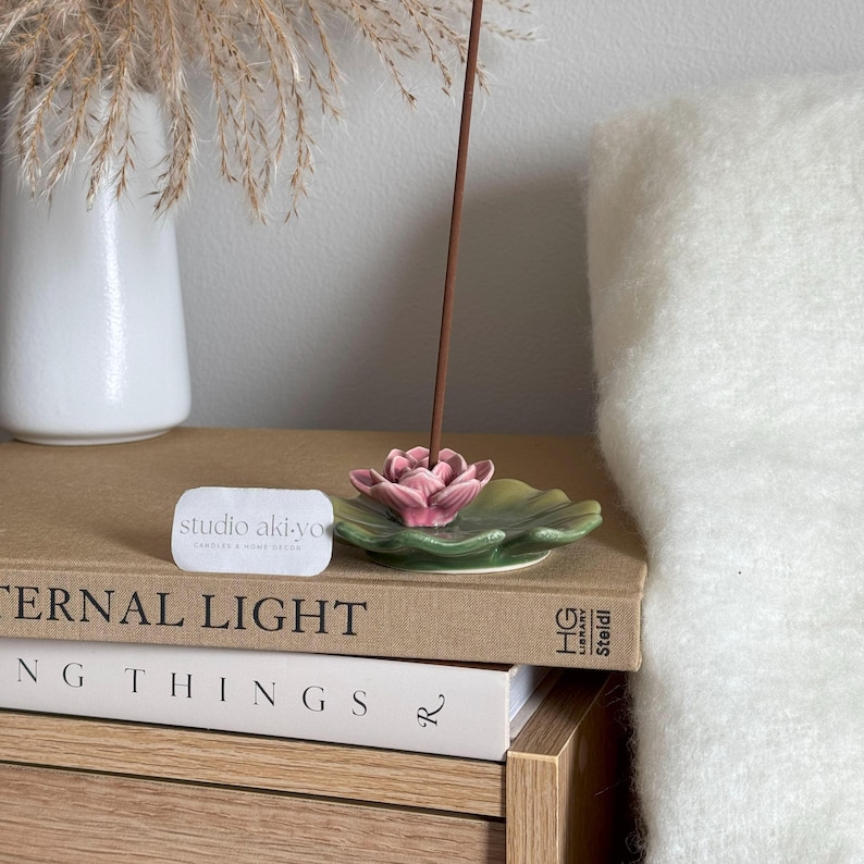 May include: A pink and green ceramic lotus flower incense holder with a brown incense stick burning. The holder is sitting on a stack of books with the text "Eternal Light" and "Studio Aki-yo Candles & Home Decor" visible.