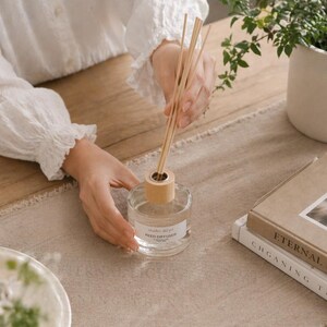 May include: A clear glass reed diffuser bottle with a light wood collar and several wooden reeds inserted into the top. The bottle features a white label with the text studio akiyo REED DIFFUSER printed in black, containing a clear liquid.