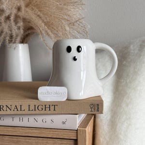 May include: A white ceramic ghost-shaped mug with black eyes and a black mouth. The mug has a handle and sits on a stack of books. A small white rectangular object with the text "studio aki-yo" is in front of the mug.