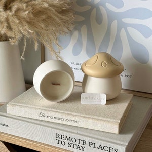 May include: Two ceramic candle holders shaped like mushrooms. The larger holder is white with a beige mushroom top. The smaller holder is white and round. Both holders are on top of two books.