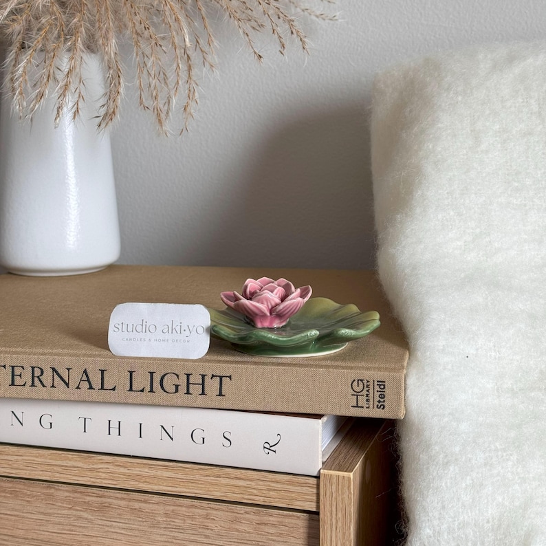 May include: A pink ceramic lotus flower sits on a green ceramic leaf dish. The dish is on top of a stack of books. The top book has the text "Eternal Light" on the cover. A business card with the text "studio aki-yo" is in the foreground.