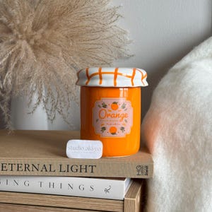 May include: Orange ceramic jar candle with a white lid and a label that says "Orange" with a small orange illustration. The jar is sitting on a stack of books.