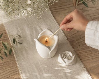 Op de afbeelding: Een witte keramische kandelaar in de vorm van een tulp, met een brandende kaars erin. Er ligt een klein, wit roosvormig deksel naast. Een lucifer wordt gebruikt om de kaars aan te steken. De scène speelt zich af op een neutraal gekleurde loper.