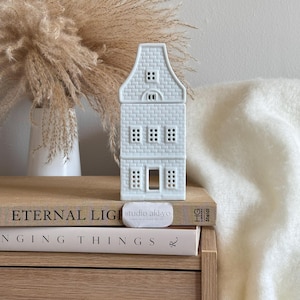 Peut inclure: Un photophore en céramique bleu clair en forme de maison avec un motif de briques. La maison a plusieurs fenêtres et une porte. Le photophore est posé sur une pile de livres. Le texte "studio aki-yo" est sur un petit objet blanc.