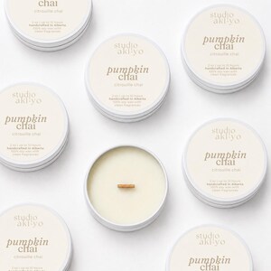 May include: Several round white metal tins containing soy wax candles with wooden wicks. Each lid features the text studio aki-yo, pumpkin chai, citrouille chai, 2 oz, up to 10 hours, handcrafted in Alberta, and 100% soy wax with clean fragrances.