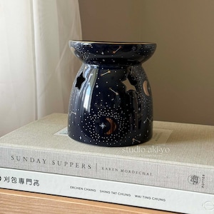 May include: A black ceramic oil burner with a gold rim and a celestial design. The design features stars, a crescent moon, and a starburst. The oil burner is sitting on a stack of books.