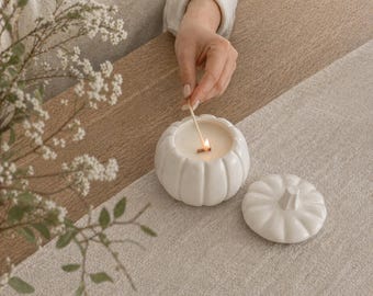 May include: A white ceramic pumpkin-shaped candle with a lit wick. The candle sits on a beige table runner. The lid of the candle is next to the candle. A hand is lighting the candle with a match. Home decor.