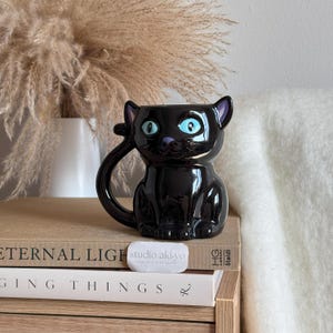 Peut inclure: Une tasse noire en forme de chat avec des yeux bleus et une poignée en forme de queue incurvée. La tasse est posée sur une pile de livres. La tasse porte le texte "studio aki-yo" sur une petite étiquette blanche.
