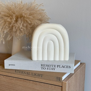 May include: White ceramic vase shaped like a rainbow arch. The vase is sitting on top of two books, one titled "Remote Places to Stay" and the other titled "Eternal Light".