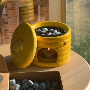 Peut inclure: Un brûleur d'huiles essentielles en céramique jaune avec une découpe en forme d'étoile et une bougie chauffe-plat allumée à l'intérieur. Le brûleur est ouvert, révélant des pierres grises dans un liquide foncé. Une boîte de fondants parfumés gris est à proximité.