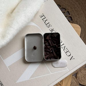 May include: A silver metal tin containing dark brown incense cones, with one cone placed separately. The tin rests on a beige surface with the text "A CENTURY OF STYLE". A small white rectangular object with text is also present.