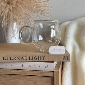 May include: A clear glass mug with a rounded, double-walled design, a curved handle, and small feet at the base. A small white rectangular tag with the text studio aki-yo is placed in front of the mug, which rests on a stack of two books.
