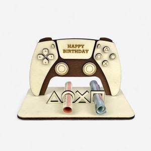 Könnte beinhalten: Ein hölzerner PlayStation-Controller-Geldhalter mit der Aufschrift "Happy Birthday" auf der Vorderseite. Der Controller hält zwei aufgerollte Geldscheine.
