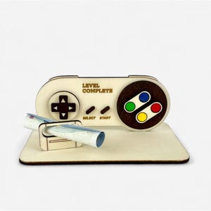 Könnte beinhalten: Ein Geschenkkartenhalter aus Holz in Form eines Retro-Videospiel-Controllers. Der Controller hat ein Steuerkreuz, Tasten und die Worte "LEVEL COMPLETE" und "SELECT START". Eine aufgerollte Banknote befindet sich im Halter.