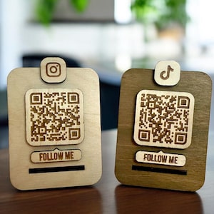 Könnte beinhalten: Zwei Holzschilder mit QR-Codes und Social-Media-Logos. Das Schild links hat ein Instagram-Logo und den Text "FOLLOW ME". Das Schild rechts hat ein TikTok-Logo und den Text "FOLLOW ME".