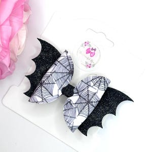 May include: A hair bow featuring black glittery bat wings on the sides and a central section with a white and black spiderweb pattern. The bow is attached to a white display card that includes a circular logo with the text The Graceful Rose.