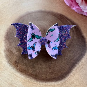 May include: A hair bow featuring a central lavender section with a repeating pattern of black, blue, and teal bats and small stars. The outer wings are made of dark purple glitter material, and a small purple glitter knot sits in the center.