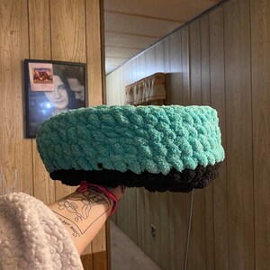 May include: A teal and black crocheted headband with a textured design. The headband is being held up by a hand with a tattoo.