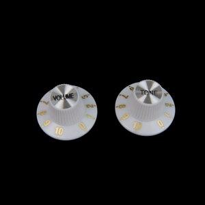 Könnte beinhalten: Zwei weiße Gitarrenknöpfe mit goldenen Zahlen und Beschriftung. Der Knopf links sagt "VOLUME" und der Knopf rechts sagt "TONE".