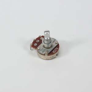 以下が含まれることがあります： 小型電子部品である可変抵抗器のクローズアップ。銀色の金属ボディ、ねじ付きシャフト、はんだ付けポイントのある茶色の回路基板を備えています。「A25K」の文字が側面に印刷されています。