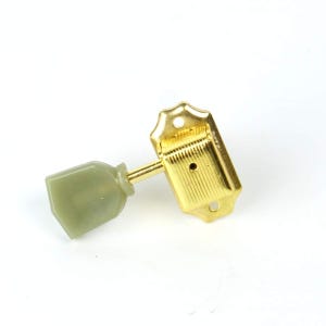 May include: A gold-colored metal guitar tuning peg featuring a rectangular base with two screw holes and a textured surface. The tuning key is made of a solid, opaque, light olive-green plastic material with a smooth, slightly tapered shape.