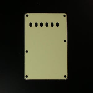 May include: A cream-colored guitar back plate with six screw holes and a rounded rectangular shape.